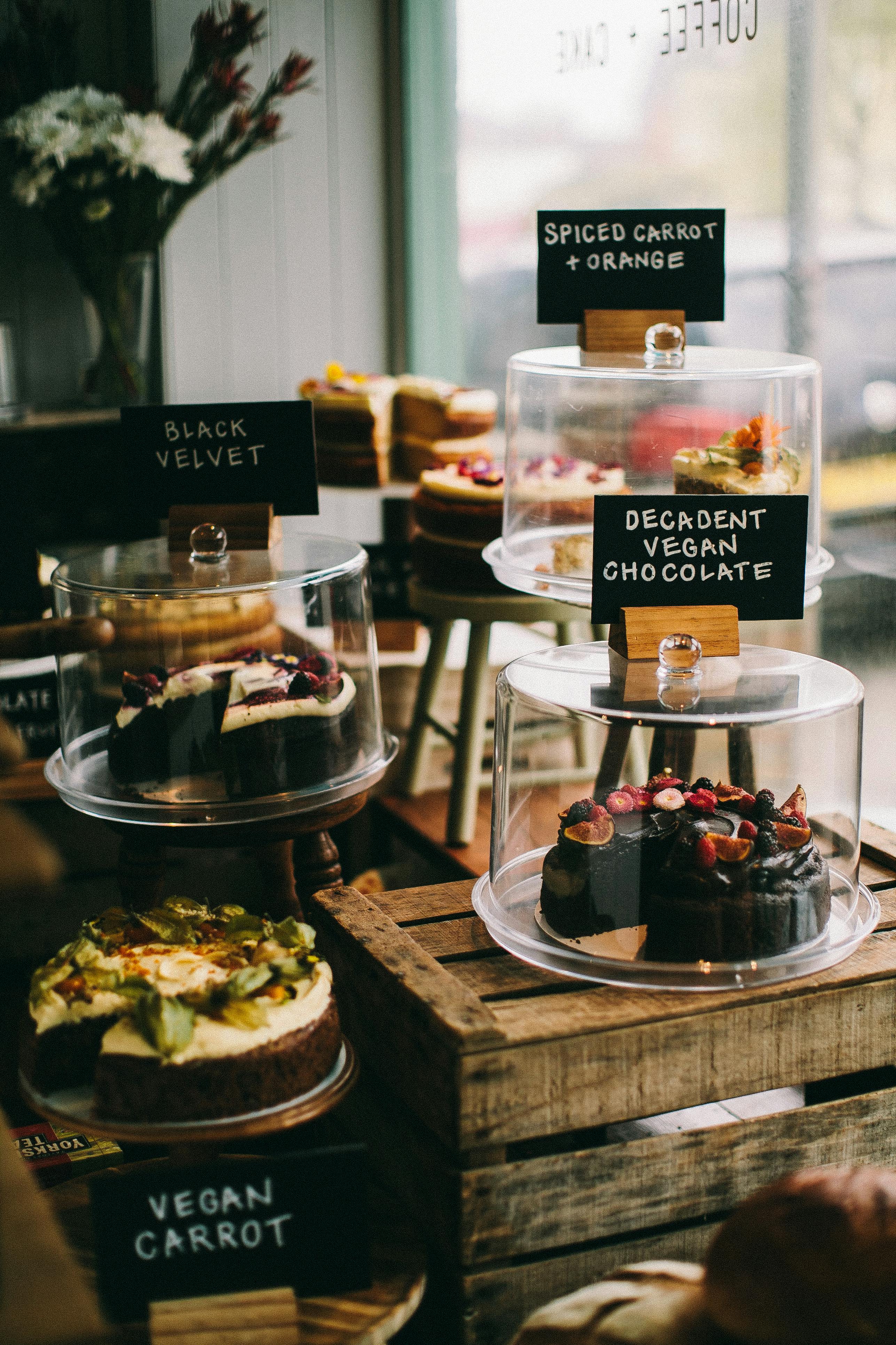 Vegan feestje organiseren: het complete stappenplan Vitrine met vegan taarten onder stolpen, waaronder chocoladetaart met vers fruit, worteltaart en black velvet cake