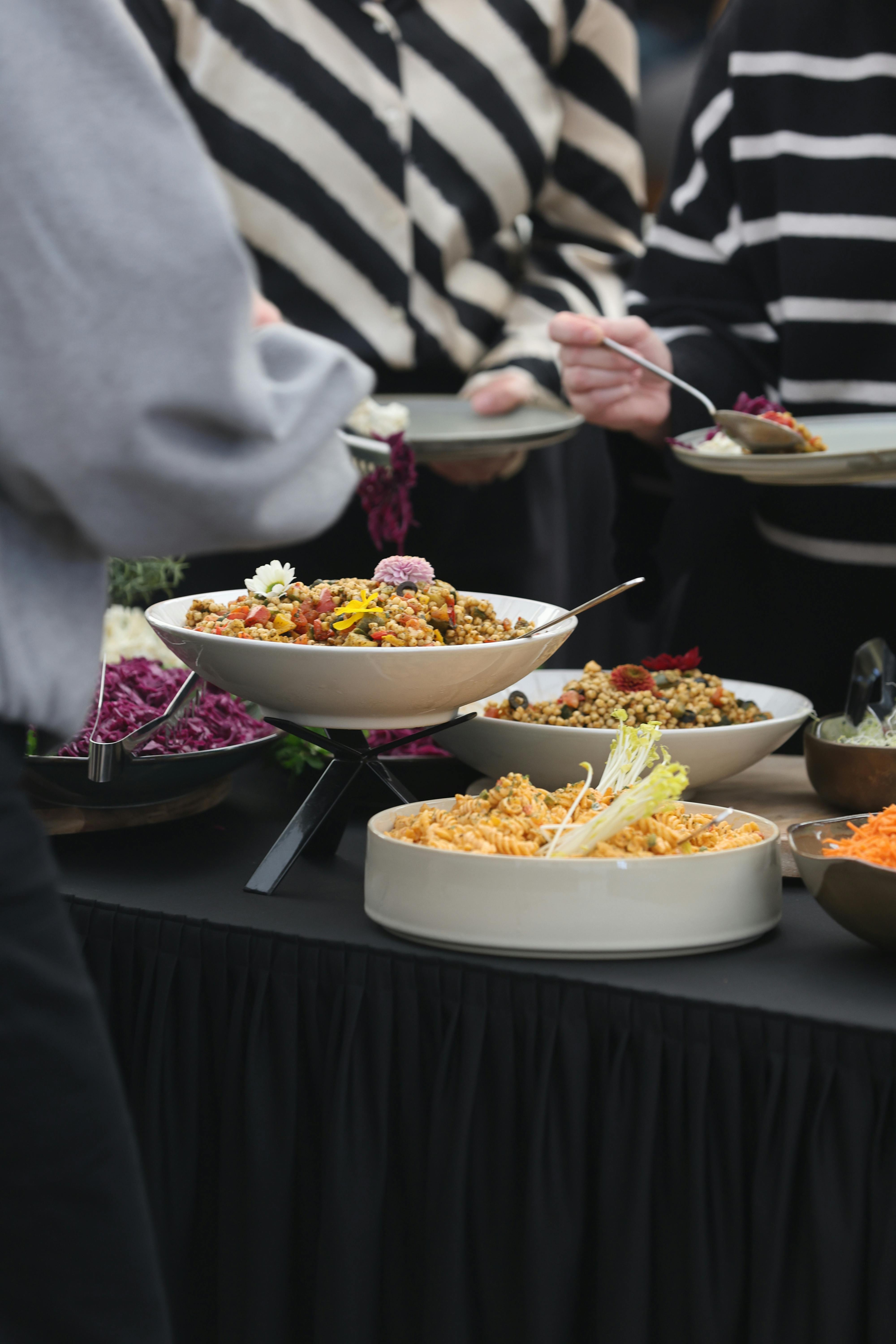 Vegan catering op het werk: van borrel tot klantevent Gasten scheppen op bij een plantaardig cateringbuffet met graansalades, pasta en groenten tijdens een buitenevenement