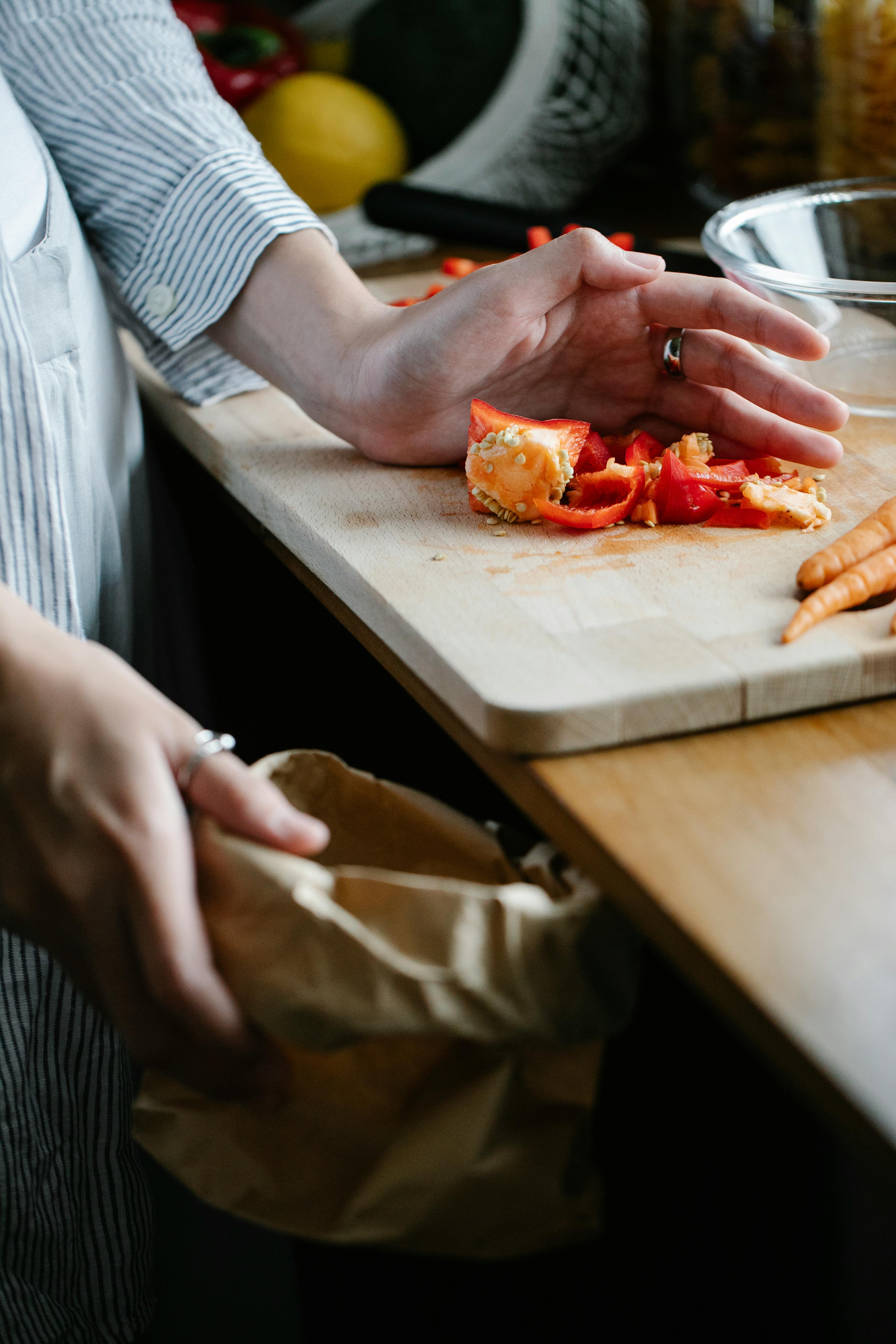 Persoon in schort verzamelt groenteafval van rode paprika in een papieren zak tijdens het koken